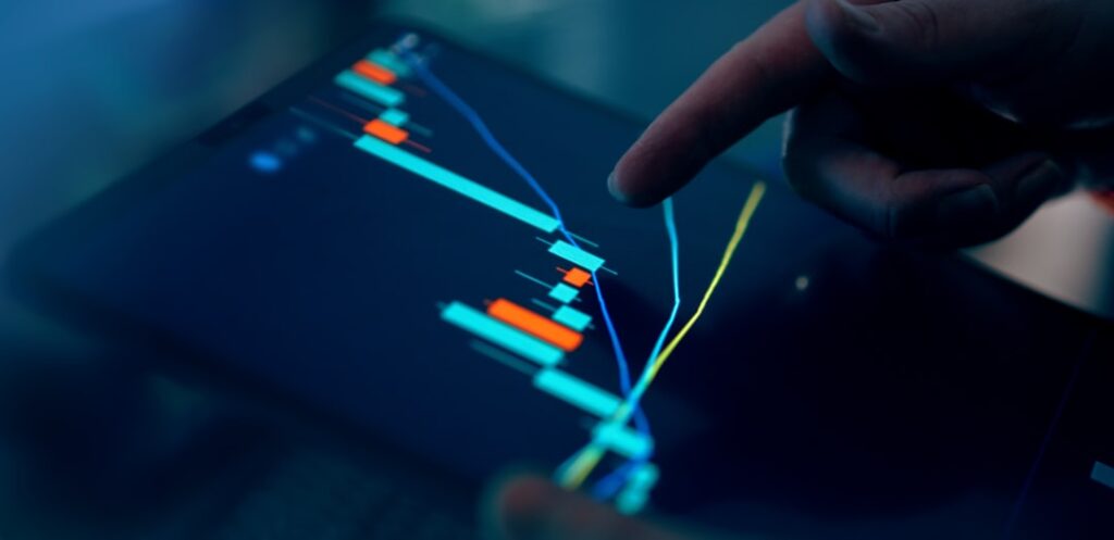 A hand pointing at a colorful trading chart on a tablet screen in a dimly lit environment.